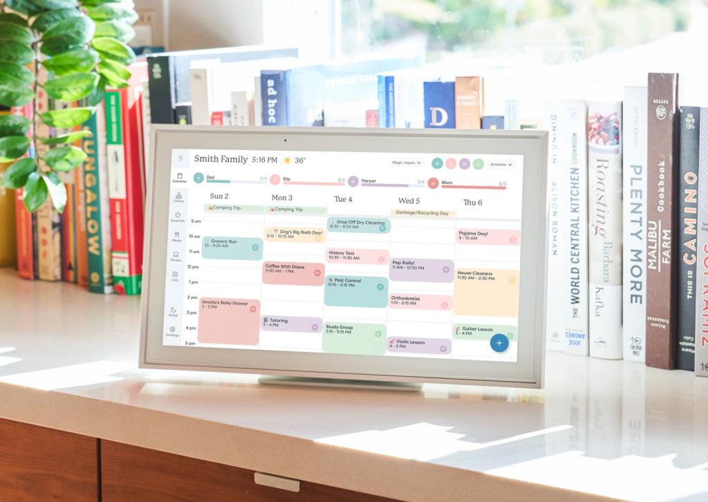 A digital family calendar displayed on a tablet, showcasing a weekly schedule with various colored events, set against a backdrop of books and a green plant.